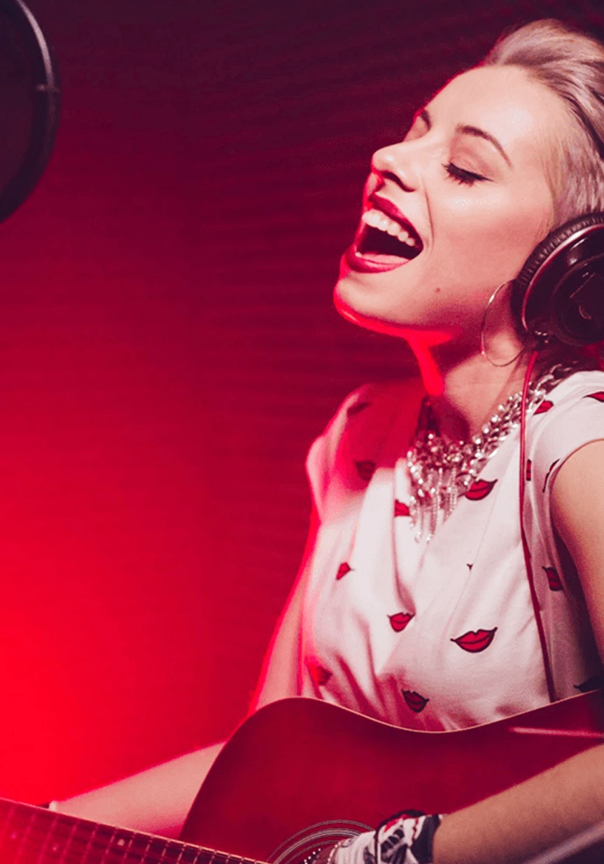A woman wearing headphones sings passionately into a microphone while playing an acoustic guitar, bathed in pinkish-red studio lighting.