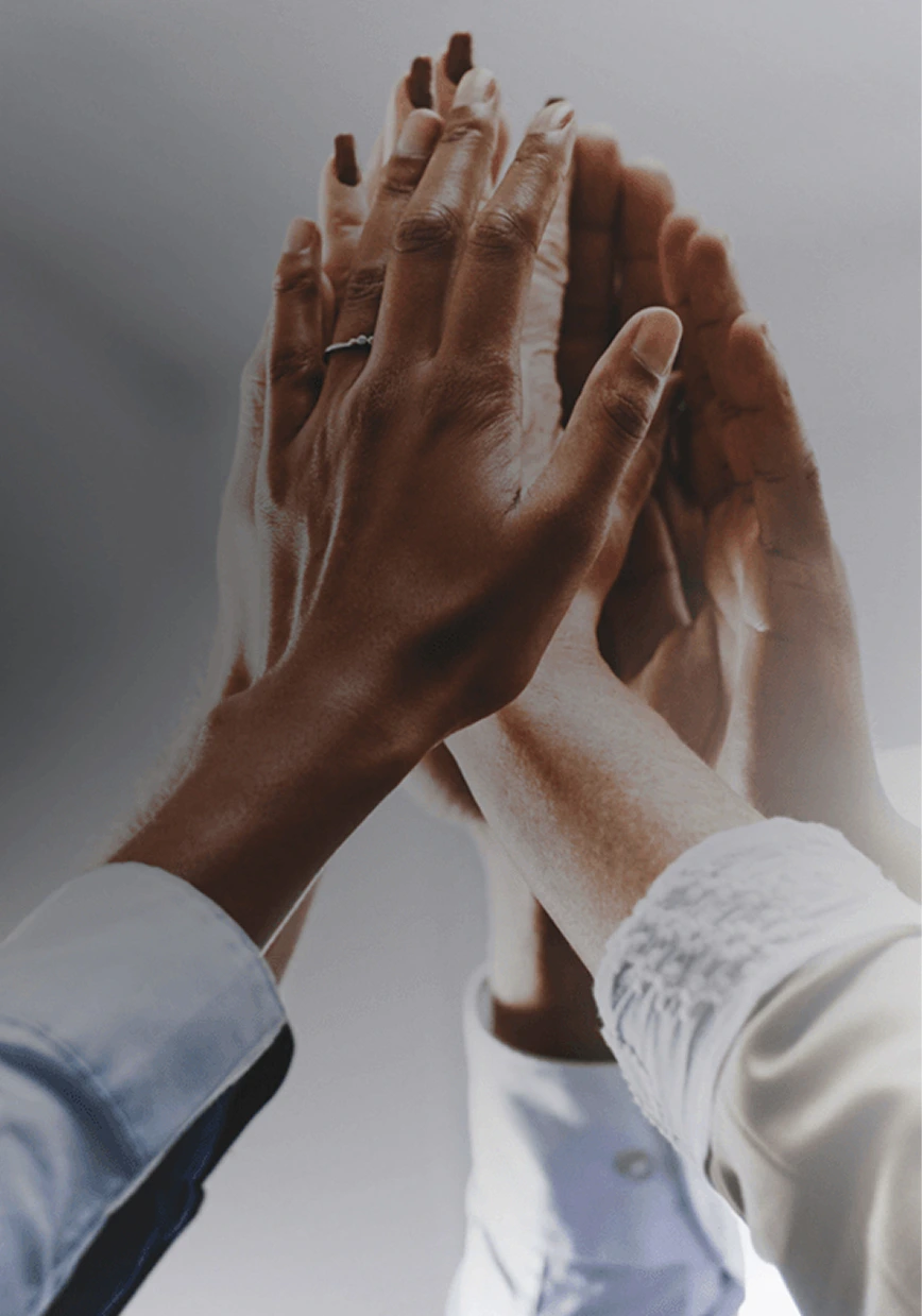 Four hands of diverse skin tones, some wearing white sleeves, come together in a high-five gesture against a blurred, gray background. One hand wears a ring. The image suggests teamwork and unity.
