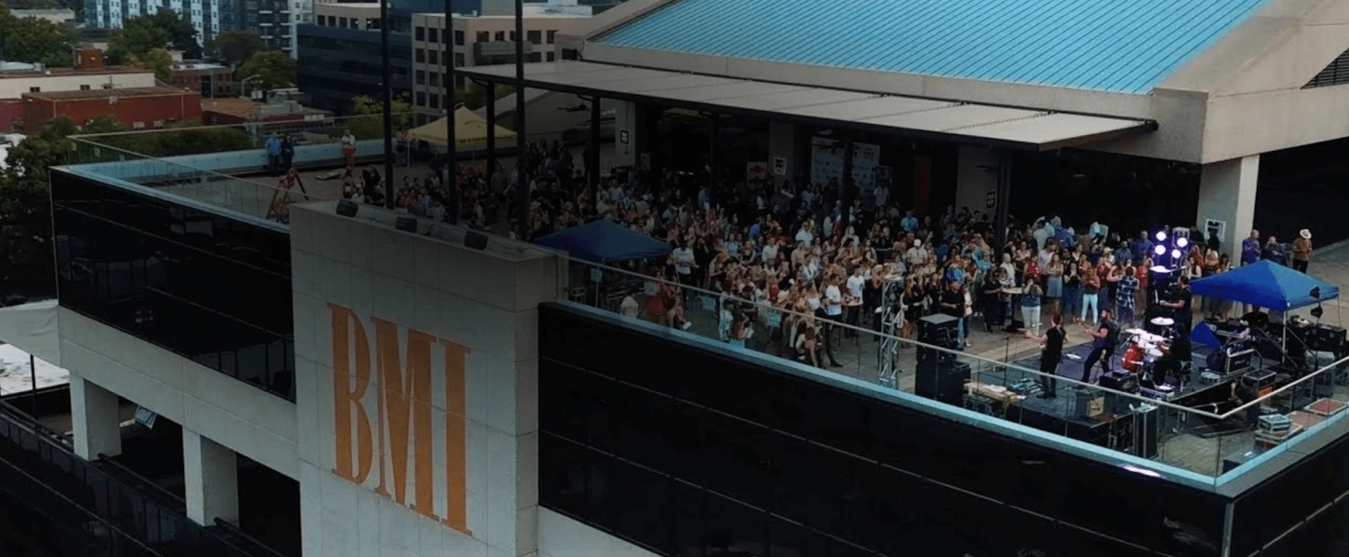 A crowd gathers on a rooftop terrace for a live music performance at the BMI building, with musicians playing under blue umbrellas and city buildings in the background.