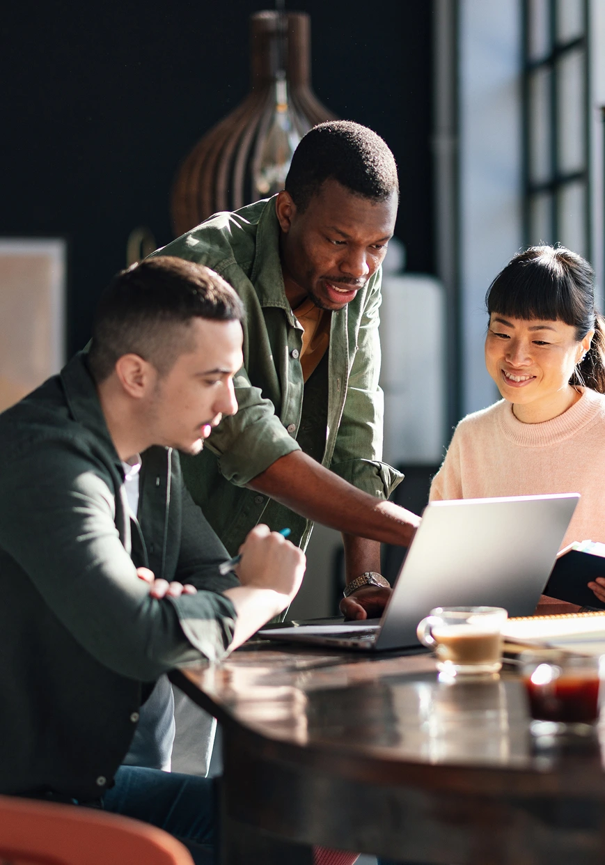 Three people sit at a round table, collaborating and looking at a laptop. One person points at the screen while the others listen and smile. Sunlight streams in through a window, creating a bright, productive atmosphere.